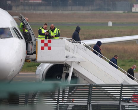 Officers stand at a plane's stairway as deported immigrants wearing hoods and masks descend