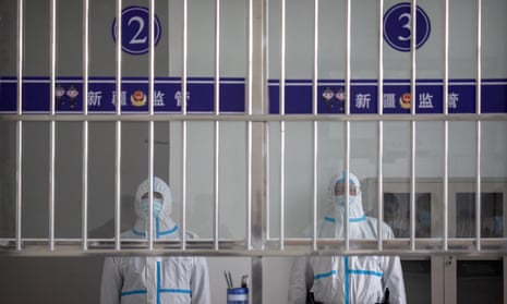 Two men at attention, standing behind bars and wearing protective suits and masks that leave only eyes showing.