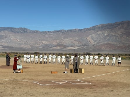 A line of baseball players in white uniforms on a dusty baseball field, with a man, a woman, and two teenage girls in period costume holding suitcases.