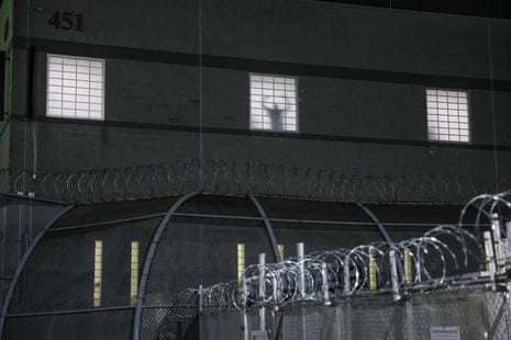 A detainee gestures through a lit window above a razor-wire fence at a detention centre