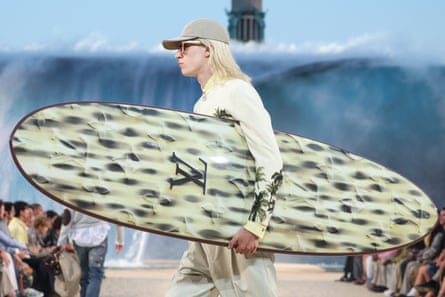 A blond male model on the catwalk carrying a surfboard with the LV logo, and wearing LV clothes and cap