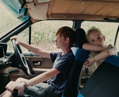A red-haired boy drives a vehicle while a blond-haired girl rests her chin on her arms in the passenger seat