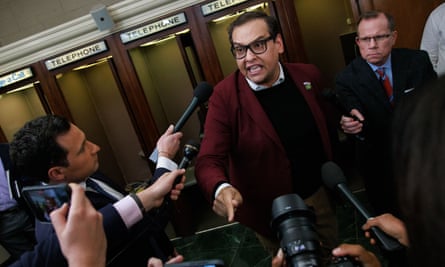 A round-iush Latino man with dark hair and dark eyeglasses, wearing a dark mauve suit jacket over a sweater and collared shirt points with one finger as he appears to snarl in the direction of multiple hands holding out cellphones and microphones.