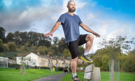 Fitness expert Joel Snape, wearing shorts and a T-shirt, standing on a bench with his arms out for balance, in a park, houses in the background. He has one foot up on the seat of the bench and the other on the back of the bench.