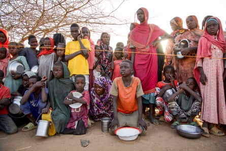 A group of Sudanese people, mainly women and children, wait with empty bowls and food containers.