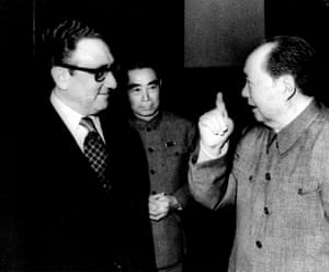 A jowly middle-aged man with a high forehead in heavy glasses wearing a tie and dark western suit (Kissinger) listens to Mao, an elderly Chinese man in a plain grey tunic, as another Chinese man stands nearby