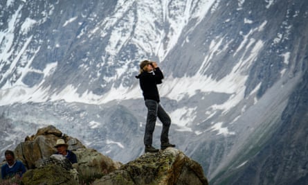 A trekker standing on top of a rock looking at a glacier through binoculars