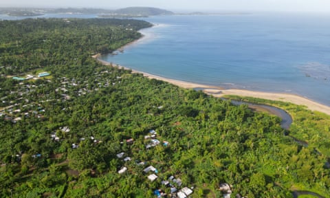 The Pacific island of Vanuatu: aerial view of lush forestation with a small, scattered settlement towards the edge of the coast and blue sea