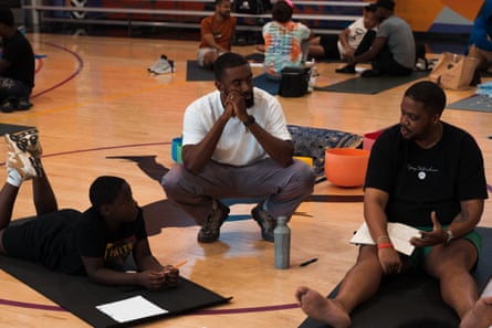A man in a white shirt squats next to a man and a child who are seated on adjacent yoga mats holding journals.
