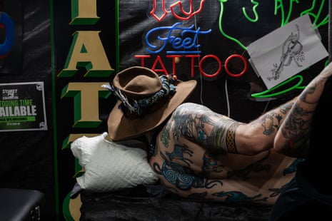 A person with a tattooed back and a wide brimmed hat lies on their side in front of a partly visible neon sign that says ‘feet tattoo’