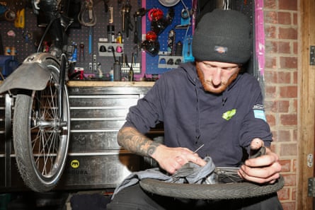 A man with a ginger beard wearing a black beanie hat and hoodie works on a bicycle wheel. He has a tattooed arm and oil on his fingers. There are tools on the wall behind him and a bicycle to his side.