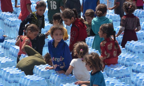 Syrian children at a refugee camp stand among stacks of water bottles