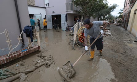 A man shovels mud and water from a street as people stand behind in a flooded yard