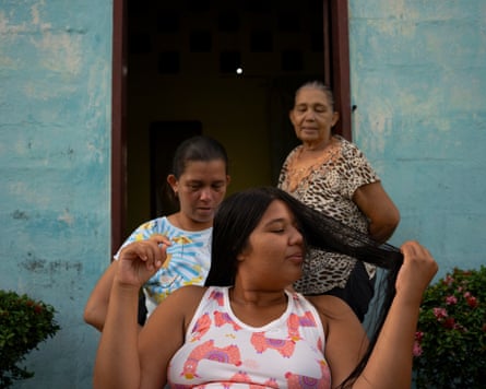 Carmen Guedez has her hair done by her aunt, Edismar García, while her grandmother, Carmen Sivira, watches from the front door of her house in Poblado Uno