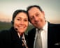 Alfredo and Carolina, who is wearing a headset, stand with their heads together, smiling, outdoors at dusk