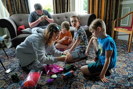 Sophia, who wears a grey hoodie, sits on a patterned carpet with three young boys as they look at craft materials and wool; an older boy sits on a grey sofa behind them.
