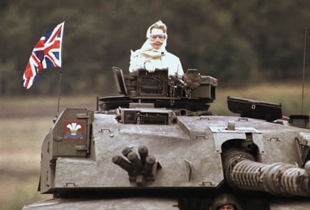 Margaret Thatcher standing in a tank with the union jack on it, wearing a cream coat, a scarf around her hair, and goggles