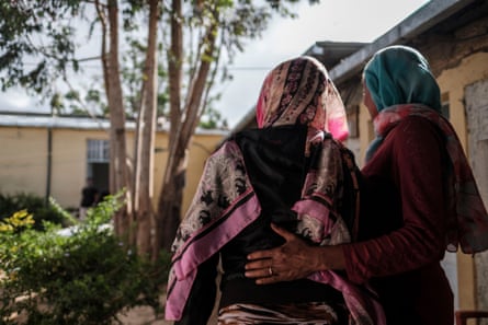 Two women stand with their backs to the camera. One has her arm around the other’s waist