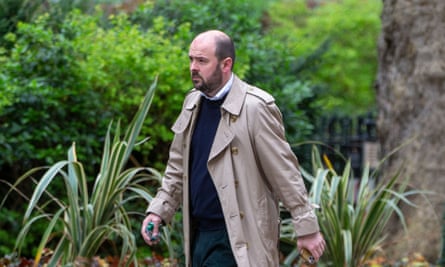 Richard Holden, in a light coloured raincoat, walking along a street