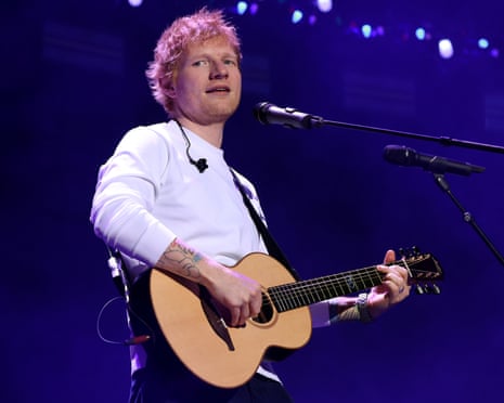 Ed Sheeran sings into a microphone while playing acoustic guitar onstage