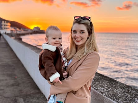 A smiling woman standing on a seafront holding a small child in her arms.