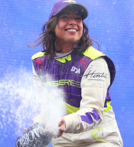Jamie Chadwick in her racing gear and cap, smiling broadly as she opens a bottle of Champagne, spraying its contents, after winning the inaugural W Series.