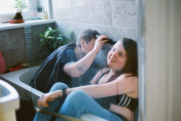 Two people sitting clothed in a bath, one cutting the other’s hair