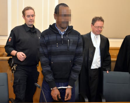 A man in handcuffs is flanked by a guard and lawyer in a German courtroom