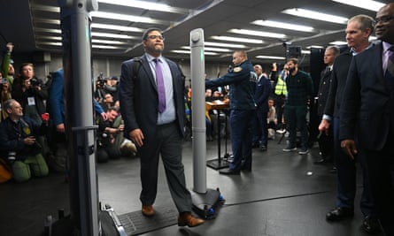 A round white-presenting man in a suit and purple tie walks through what is sort of like an airport scanner or metal detector, with two gray pillars making a sort of doorway.