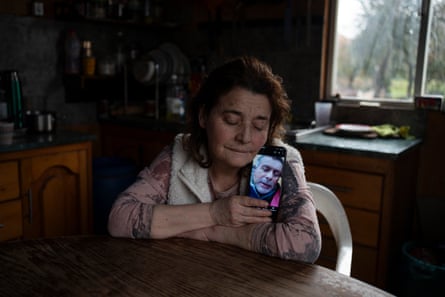 A woman sitting at a table in a kitchen holds a phone next to her head, the screen showing a photograph of a man’s head. Her eyes are closed