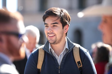 Young white man with brown hair and light beard, smiling outdoors, wearing backpack, in collared shirt and fleece.