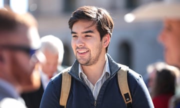 Young white man with brown hair and light beard, smiling outdoors, wearing backpack, in collared shirt and fleece.