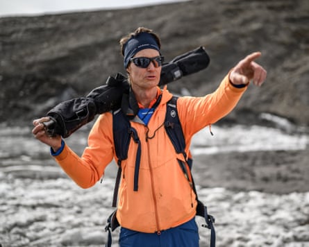 Glaciologist Matthias Huss in bright orange jacket, sunglasses, headband, pointing towards a mountain
