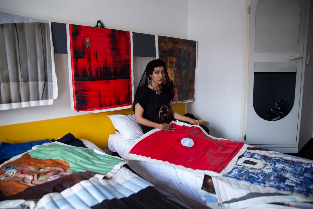 Atefeh Amini Afghan multidisciplinary artist, sits on a bed covered with brightly-covered paintings.