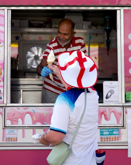 An Asian man hands a cone of vanilla ice-cream to a man in an England football top, wearing a plastic cowboy hat with the England flag on it
