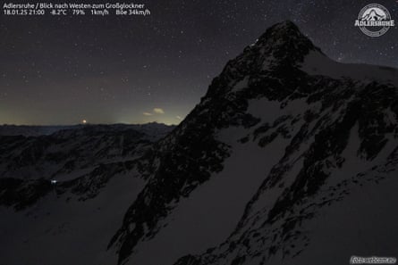 A mountain covered in snow at dusk with stars in the sky and the time and date top left on the screen.