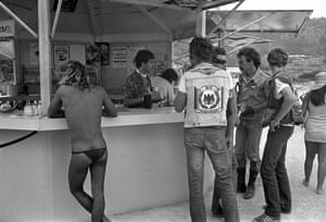 Black-and-white photo. People pictured from behind while standing at a bar outside, three men in biker jackets and jeans and one in swimming briefs