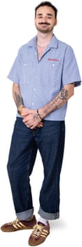 A full-length portrait of a Joe, a young man wearing jeans, trainers and a short-sleeve striped shirt, with a moustache and tattoos