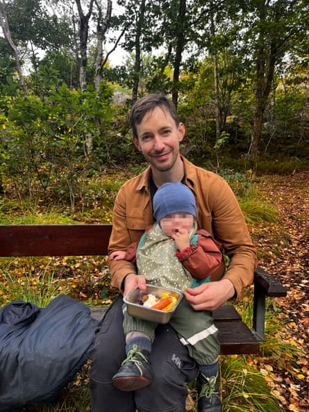 Simon Montgomery sits on a bench holding his toddler and a container of fruit in woodland