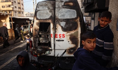 Two children stand on a street next to a burned-out white van with the remains of the word 'press' written on it in red.