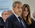 Nicolas Sarkozy looking serious and weary with his wife in the background