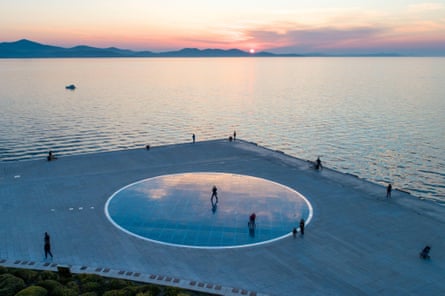 People standing around an outside art installation beside the sea at sunset.