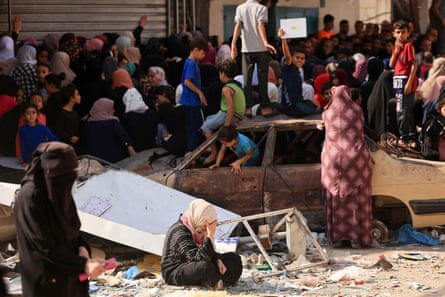 Children play in a charred car as people queue for bread in front of a bakery that was partly destroyed in an Israeli strike