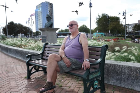 An older man in a vest and shorts sits on a park bench in front of a flowerbed