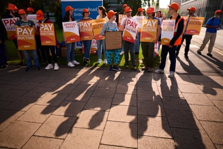 Doctors on a picket line wearing orange BMA hats and holding signs and placards; they stand in the sun and their shadows are projected in front of them.