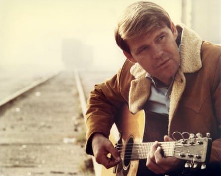 Glen Campbell, wearing a flyer jacket, strums a guitar alongside a railroad track in 1967.