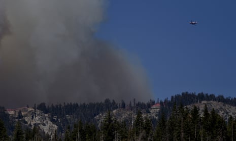 Gray smoke billows above a mountain range as an aircraft flies in the direction of the column of smoke.