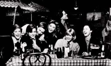 A black-and-white image of seven young men and women seated and standing at a bistro table with a checked tablecloth and roundback chairs, laughing uproariously.