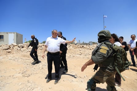 Gilad Kariv, a large man in a white shirt, stands among military personnel on a sandy site with one hand held out.