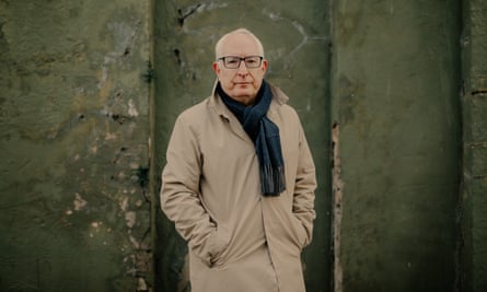 Neil Duncan-Jordan stands against a green, mossy wall in Poole. He is in his 60s and wears a black scarf and beige mac.